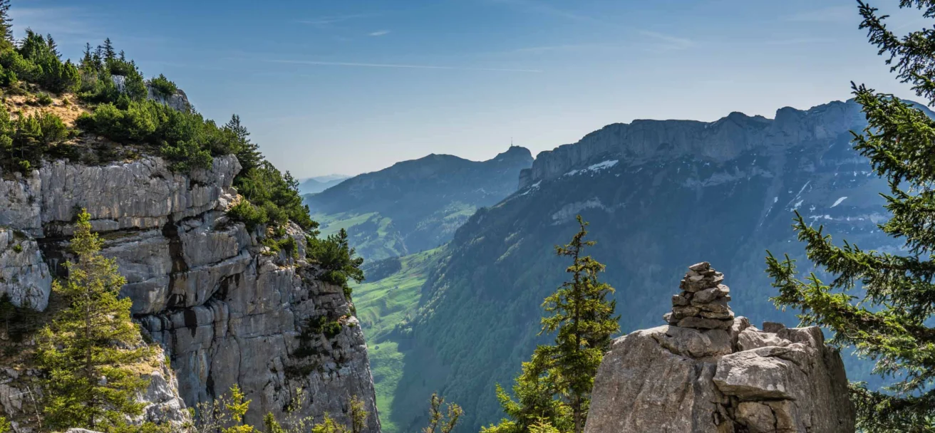 Mountain landscape with rocky cliffs, scattered trees, and a distant view of rolling hills under a clear blue sky.